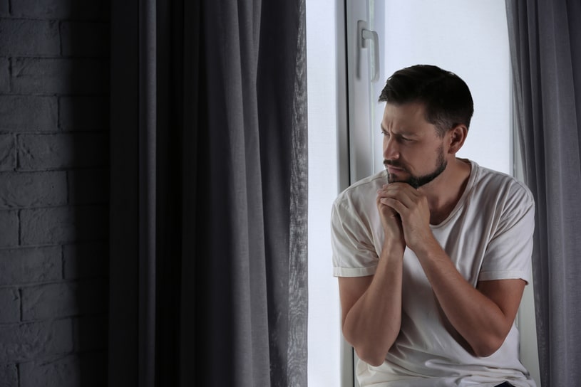 A sad man sits on the windowsill and looks sadly out the window.