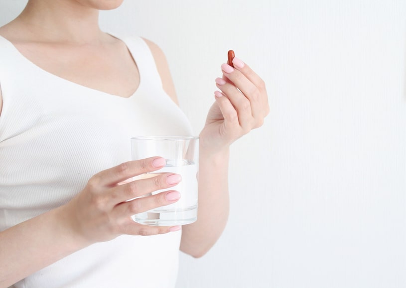 A woman in a white T-shirt holds a pill in one hand and a glass of water in the other.
