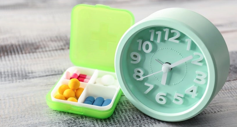 Plastic container with pills and clock on wooden table.