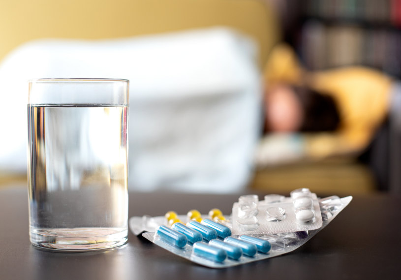 A glass of water and some pills on the table with a woman lying in bed in the background.