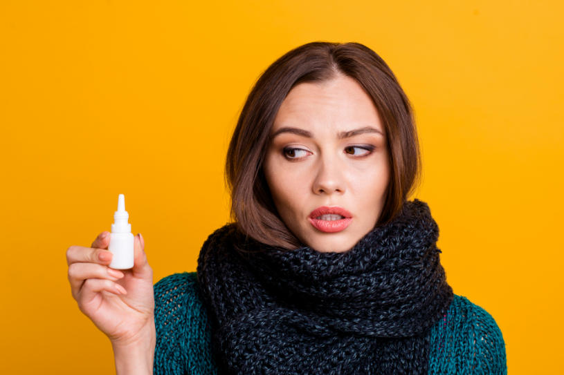 A sick woman looks at nasal spray in her hand over yellow background.