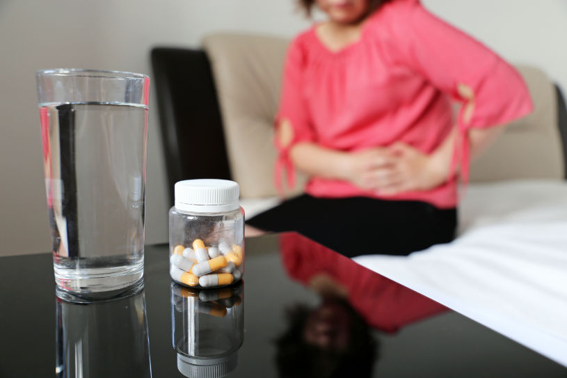 Some pills and a glass of water with a woman in pain in the background.